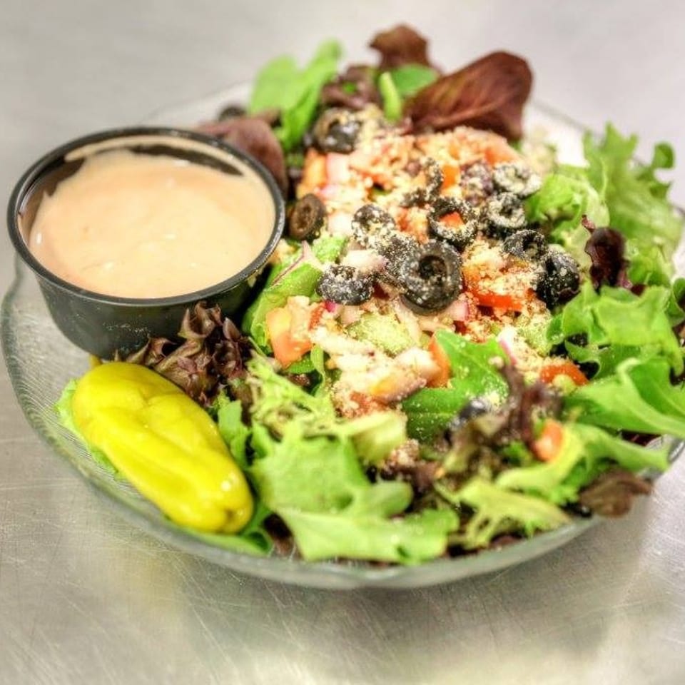 Fresh garden salad with mixed greens, tomatoes, olives, cheese, a pepperoncini, and dressing.