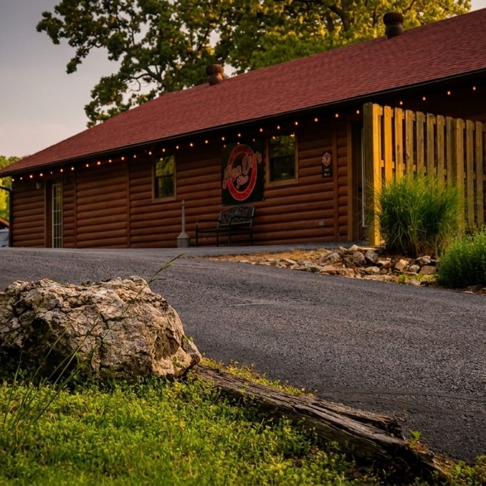 Two rustic log cabin buildings with red roofs, surrounded by green grass and trees under a warm sky.